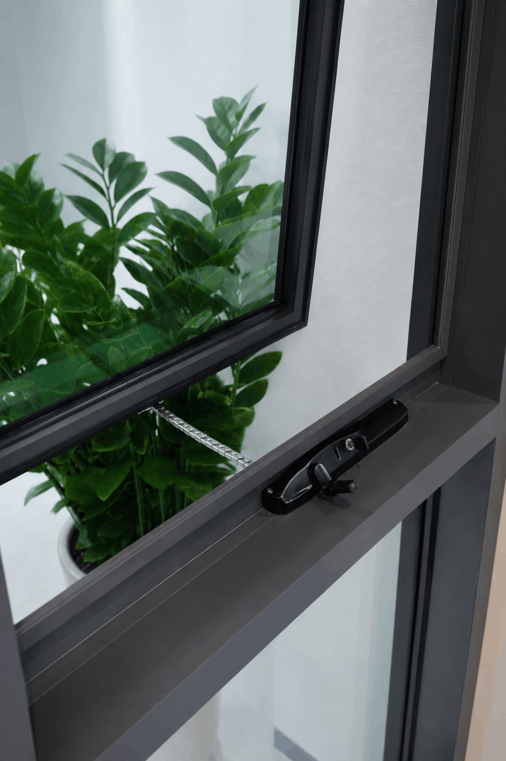 Close-up of a modern dark aluminium awning window, partially open, featuring a sleek handle and frame with green plants visible behind the glass.