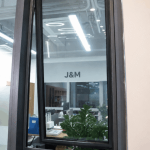 Close-up of a modern black aluminium awning window with glass panels, viewed from outside, showing an office interior and green plants behind the window.